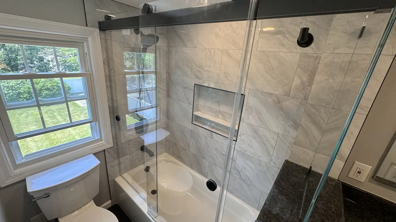 A remodeled bathroom with a glass sliding shower door, white marble-look tile and a black granite vanity